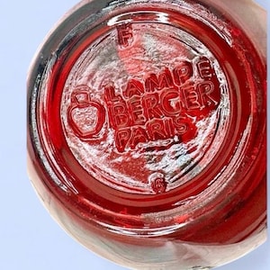 May include: A circular red glass base of a Lampe Berger Paris fragrance lamp. The underside features embossed text reading LAMPE BERGER PARIS, a small bottle icon, the letter F, and the number 6, all molded into the translucent red glass surface.