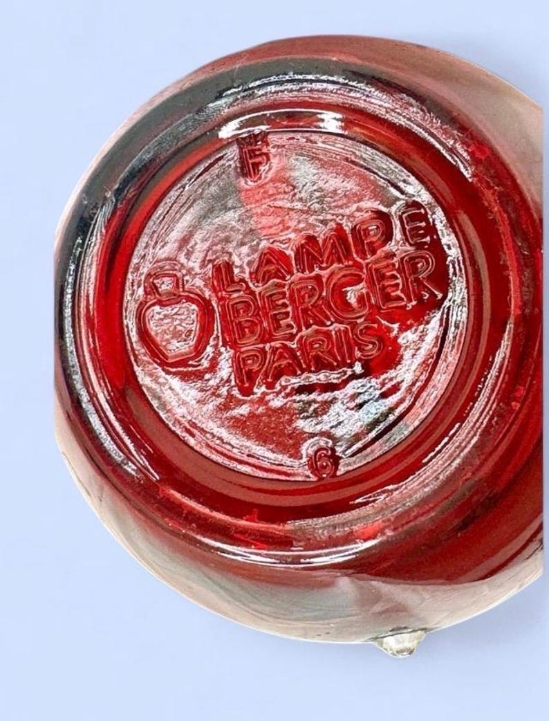 May include: A circular red glass base of a Lampe Berger Paris fragrance lamp. The underside features embossed text reading LAMPE BERGER PARIS, a small bottle icon, the letter F, and the number 6, all molded into the translucent red glass surface.