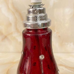 May include: A deep red glass bottle featuring a silver-toned metal neck with a stepped, tiered design. The metal collar is embossed with the text BEBERG and accented with small clear crystals. Additional clear crystals are embedded in the glass.