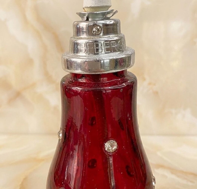 May include: A deep red glass bottle featuring a silver-toned metal neck with a stepped, tiered design. The metal collar is embossed with the text BEBERG and accented with small clear crystals. Additional clear crystals are embedded in the glass.
