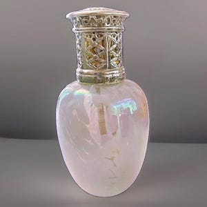 May include: A pearlescent glass fragrance lamp with an iridescent finish that reflects light in soft pink and blue tones. The vessel features a rounded body and is topped with a decorative, perforated silver-colored metal crown cap.