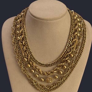 Puede incluir: Collar dorado de múltiples hilos exhibido en un maniquí. El collar presenta varios estilos de cadena, incluyendo cadenas de eslabones, hilos de cuentas y un delicado colgante de bola de filigrana. Una pieza llamativa.
