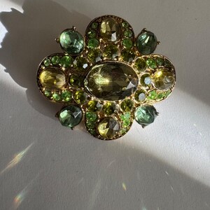 Peut inclure: Broche dor&eacute;e au design complexe. Elle pr&eacute;sente une pierre centrale ovale, entour&eacute;e de pierres pr&eacute;cieuses vertes et jaunes. Les pierres sont dispos&eacute;es en motif floral ou tr&egrave;fle, cr&eacute;ant un accessoire d&eacute;coratif.