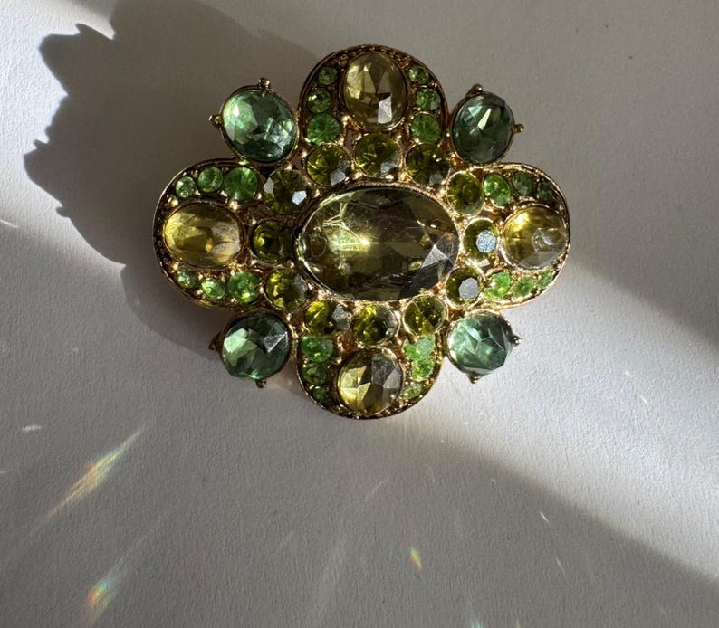 Peut inclure: Broche dor&eacute;e au design complexe. Elle pr&eacute;sente une pierre centrale ovale, entour&eacute;e de pierres pr&eacute;cieuses vertes et jaunes. Les pierres sont dispos&eacute;es en motif floral ou tr&egrave;fle, cr&eacute;ant un accessoire d&eacute;coratif.