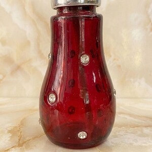 May include: A deep red glass bottle with a rounded, tapered body and a silver-toned metal cap. The exterior surface of the red glass is decorated with small, clear, circular faceted crystals arranged in a scattered pattern across the front.