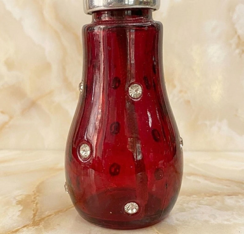 May include: A deep red glass bottle with a rounded, tapered body and a silver-toned metal cap. The exterior surface of the red glass is decorated with small, clear, circular faceted crystals arranged in a scattered pattern across the front.