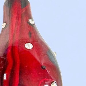 May include: A teardrop-shaped glass ornament in a deep translucent red hue, featuring a smooth, glossy surface embedded with small, clear, faceted crystals. The interior displays a darker red, elongated, organic shape visible through the glass.