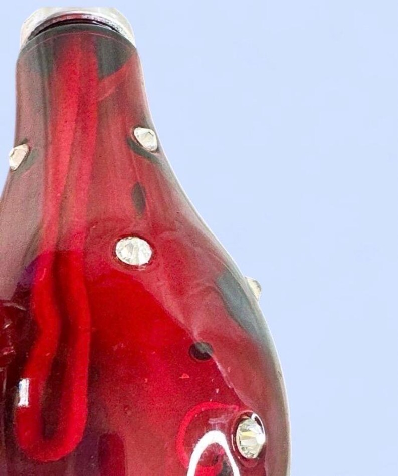 May include: A teardrop-shaped glass ornament in a deep translucent red hue, featuring a smooth, glossy surface embedded with small, clear, faceted crystals. The interior displays a darker red, elongated, organic shape visible through the glass.