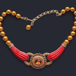 Puede incluir: Un collar dorado con cuentas grandes y redondas y un colgante central. El colgante presenta una piedra grande, ovalada, color ámbar, rodeada de piedras más pequeñas y brillantes. Acentos rojos y acanalados conectan el colgante con la cadena de cuentas.