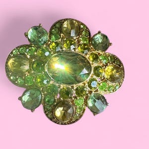 Peut inclure: Broche dor&eacute;e de forme florale orn&eacute;e d'un assortiment de pierres pr&eacute;cieuses vertes et jaunes. La broche pr&eacute;sente une pierre centrale ovale entour&eacute;e de petites pierres rondes et ovales de diff&eacute;rentes nuances de vert et de jaune.