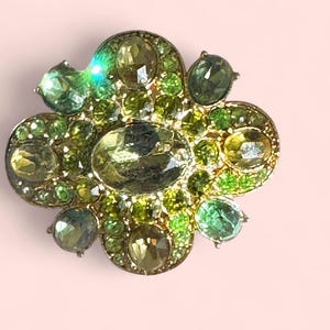 Peut inclure: Broche en forme de fleur avec une base en m&eacute;tal dor&eacute;, orn&eacute;e de pierres pr&eacute;cieuses vertes et jaune-vert de diff&eacute;rentes tailles. La broche a un design en grappe avec un m&eacute;lange de pierres rondes et ovales, cr&eacute;ant un effet scintillant. Le fond est rose clair.