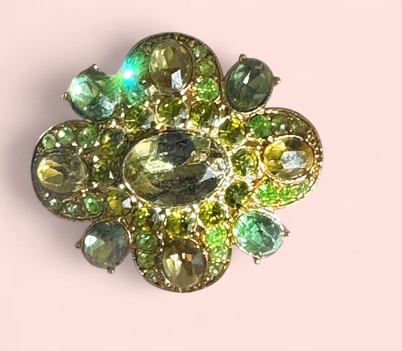 Peut inclure: Broche en forme de fleur avec une base en m&eacute;tal dor&eacute;, orn&eacute;e de pierres pr&eacute;cieuses vertes et jaune-vert de diff&eacute;rentes tailles. La broche a un design en grappe avec un m&eacute;lange de pierres rondes et ovales, cr&eacute;ant un effet scintillant. Le fond est rose clair.
