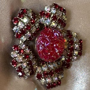 May include: A floral-shaped brooch featuring a central circular red cabochon with a cracked texture, surrounded by petals encrusted with alternating rows of small, sparkling red and clear rhinestones set in a silver-toned metal frame.