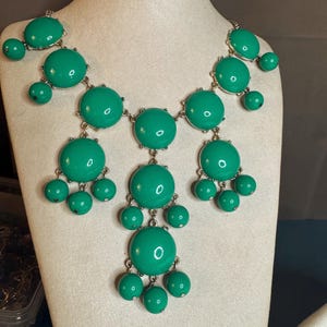 May include: A statement necklace featuring large, round, glossy, emerald green pendants and smaller matching beads. The necklace has a silver-tone metal chain and is displayed on a mannequin. The design is a bold, eye-catching piece of jewelry.