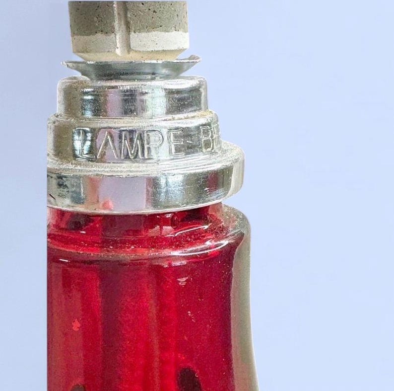 May include: A red glass lamp base featuring a silver-toned metal collar with the embossed text LAMPE B. The top of the lamp includes a grey stone-like burner component with a central vertical slit, set into a tiered metallic neck structure.