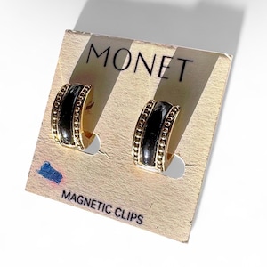 May include: A pair of gold-toned hoop earrings with black enamel centers, displayed on a Monet branded card. The earrings have a beaded edge and are labeled as magnetic clips.