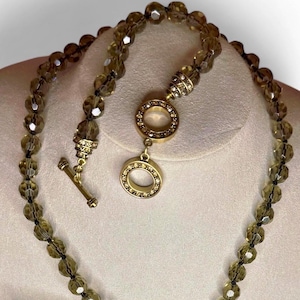 Può includere: Una collana di perline con perline sfaccettate color quarzo fumé. La collana ha una chiusura e elementi decorativi color oro, tra cui un ciondolo circolare con cristalli incastonati. Le perline sono infilate su un cordoncino scuro.