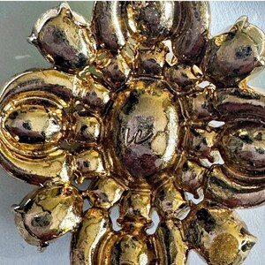 Peut inclure: Gros plan sur une broche dor&eacute;e avec un motif floral. La broche pr&eacute;sente un &eacute;l&eacute;ment central avec la lettre "W" et est entour&eacute;e de formes en forme de p&eacute;tales. Le m&eacute;tal a une surface brillante et r&eacute;fl&eacute;chissante.