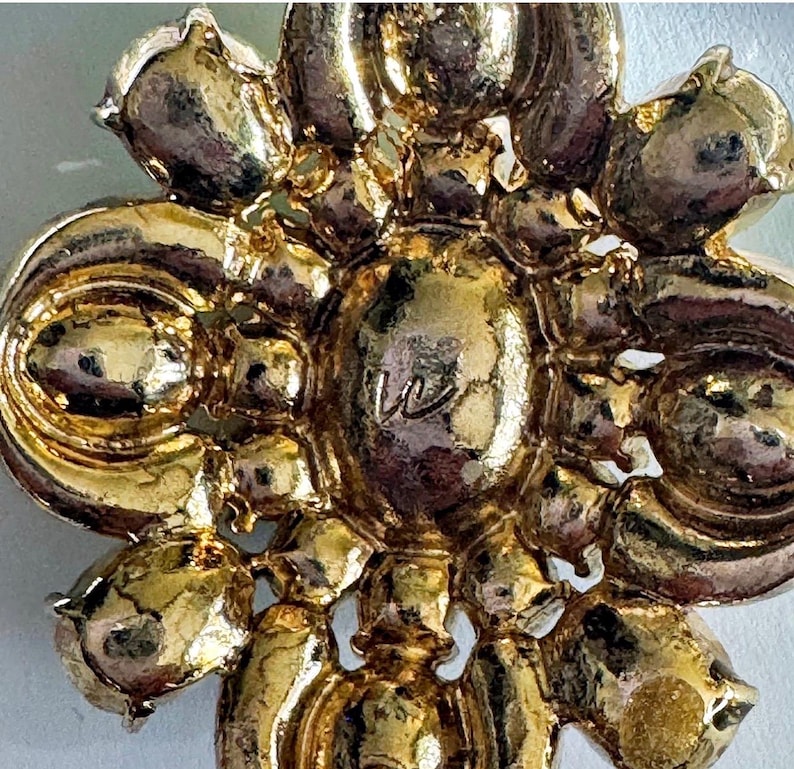 Peut inclure: Gros plan sur une broche dor&eacute;e avec un motif floral. La broche pr&eacute;sente un &eacute;l&eacute;ment central avec la lettre "W" et est entour&eacute;e de formes en forme de p&eacute;tales. Le m&eacute;tal a une surface brillante et r&eacute;fl&eacute;chissante.