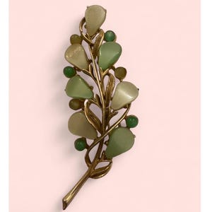 May include: A vintage gold-tone brooch in the shape of a stylized leaf or branch. The brooch features teardrop-shaped and round green and off-white stones. The design is reminiscent of a botanical theme, suitable for adding a touch of nature-inspired elegance.