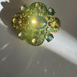 Peut inclure: Broche dor&eacute;e de forme florale, orn&eacute;e de pierres pr&eacute;cieuses vertes de diff&eacute;rentes nuances. La broche pr&eacute;sente une pierre centrale ovale, entour&eacute;e de petits gemmes ronds et ovales. Le design est orn&eacute; et attrayant.
