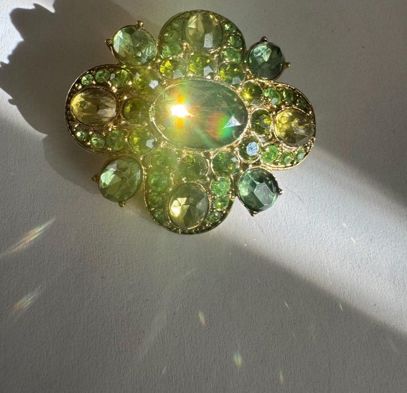 Peut inclure: Broche dor&eacute;e de forme florale, orn&eacute;e de pierres pr&eacute;cieuses vertes de diff&eacute;rentes nuances. La broche pr&eacute;sente une pierre centrale ovale, entour&eacute;e de petits gemmes ronds et ovales. Le design est orn&eacute; et attrayant.