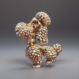 Op de afbeelding: Een goudkleurige metalen broche in de vorm van een poedel met een textuur van krullende vacht. De poedel staat in een staande houding met de kop omhoog gericht. Een kleine, enkele rode edelsteen is gezet in het oog van de poedel, wat een subtiel kleuraccent toevoegt aan de metalen afwerking van het sieraad.