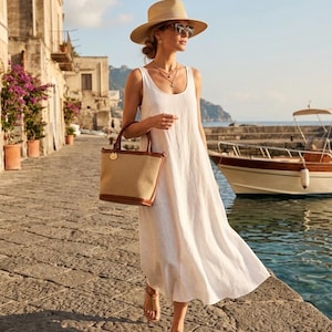 May include: A woman wearing a white linen dress, straw hat, and sunglasses walks along a stone path. She carries a beige tote bag with brown leather trim. The background features a coastal scene with buildings, water, and a boat.