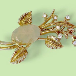 May include: A gold-toned brooch in a floral design, featuring a central oval-shaped stone and accents of clear crystals. The brooch has detailed leaves and branches, set against a light green background.