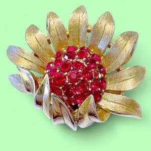 Op de afbeelding: Goudkleurige bloesembroche met een cluster van rode strass-steentjes in het midden. De broche heeft getextureerde gouden bloemblaadjes en zilverkleurige accenten, wat een opvallend contrast creëert. Dit vintage-geïnspireerde accessoire is perfect.