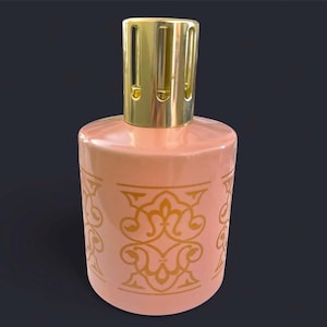 May include: A peach-colored ceramic fragrance lamp with a gold-colored metal top. The lamp features a repeating gold-colored ornate pattern around the body. The top has a cylindrical shape with vertical slits.