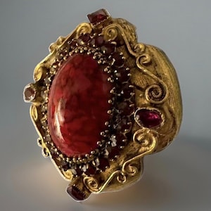 May include: A gold-toned ring featuring a large, oval-shaped red stone at the center, surrounded by a border of smaller, dark red faceted stones. The metal setting displays intricate scrollwork and textured details throughout the frame.