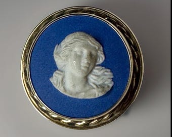 Può includere: Una spilla rotonda caratterizzata da un disco blu centrale con un rilievo bianco scolpito raffigurante un profilo umano. Il pezzo circolare è racchiuso in una cornice di metallo color oro lucido con una trama a corda ritorta sul bordo.