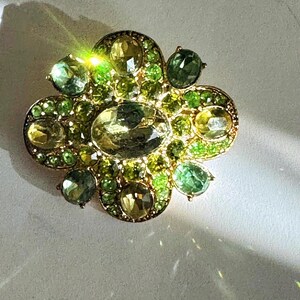 Peut inclure: Broche dor&eacute;e de forme florale orn&eacute;e de pierres pr&eacute;cieuses vertes et jaunes. La broche pr&eacute;sente une pierre centrale ovale entour&eacute;e de petites pierres rondes de nuances vertes et jaunes. Les pierres sont serties dans un m&eacute;tal dor&eacute;.