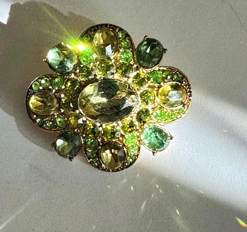 Peut inclure: Broche dor&eacute;e de forme florale orn&eacute;e de pierres pr&eacute;cieuses vertes et jaunes. La broche pr&eacute;sente une pierre centrale ovale entour&eacute;e de petites pierres rondes de nuances vertes et jaunes. Les pierres sont serties dans un m&eacute;tal dor&eacute;.