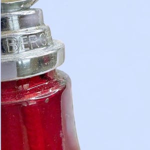 May include: A red glass vessel with a tiered, metallic silver-colored neck and cap. The metallic component features the embossed text BERGE. The glass body is translucent and deep red, showing a vertical internal structure through the glass.