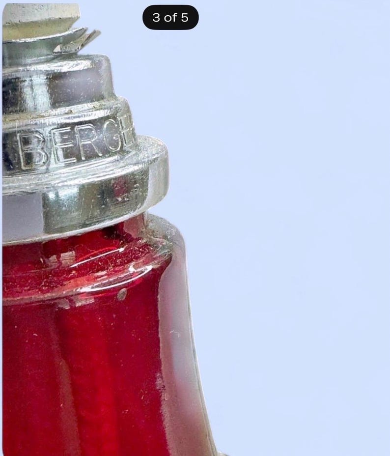 May include: A red glass vessel with a tiered, metallic silver-colored neck and cap. The metallic component features the embossed text BERGE. The glass body is translucent and deep red, showing a vertical internal structure through the glass.