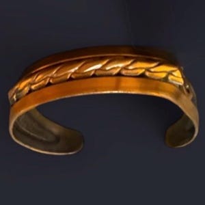 May include: A gold-colored cuff bracelet with an open design. The bracelet features a detailed leaf pattern along the top edge. The metal has a polished finish, reflecting light. The bracelet is designed to be worn on the wrist.