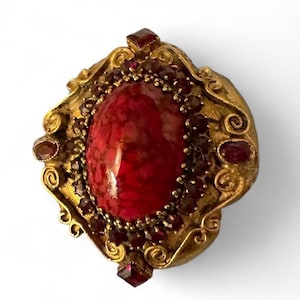 Puede incluir: Broche dorado ornamentado con una gran gema roja ovalada en el centro. La gema está rodeada de piedras rojas más pequeñas y un marco dorado decorativo. El broche tiene una estética antigua o vintage.