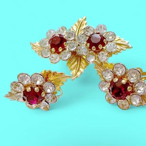 May include: A vintage jewelry set featuring a brooch and a pair of earrings. Each piece has a gold-tone metal floral design with a central red gemstone surrounded by clear crystals. The set is displayed on a turquoise background.