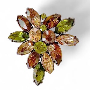 Puede incluir: Broche floral con piedras preciosas facetadas en tonos ámbar, verde oliva y topacio. El broche tiene un engaste de metal plateado y un cierre de alfiler. El diseño recuerda a una flor o estrella estilizada.
