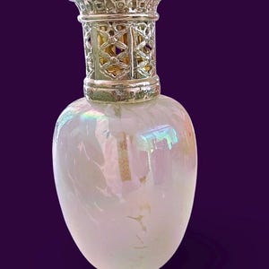 Könnte beinhalten: Eine perlmuttweiße Glas-Duftlampe mit silbernem Filigranaufsatz. Die Lampe hat einen ovalen Körper und eine dekorative silberne Kappe. Die Oberseite weist ein kompliziertes Gittermuster auf. Die Lampe steht vor einem dunkelvioletten Hintergrund.