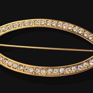 May include: An oval-shaped gold-tone brooch featuring a series of small, clear, round rhinestones set along the entire frame. The brooch is accented with a single, larger, clear, trapezoidal-shaped stone at each of the two pointed ends.