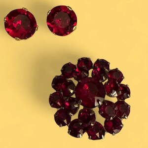 Peut inclure: Un ensemble de bijoux vintage comprenant un broche et des boucles d'oreilles assorties. La broche est un motif floral avec une grosse pierre rouge foncé ronde au centre, entourée de pierres plus petites. Les boucles d'oreilles sont rondes, avec une seule pierre rouge foncé chacune.