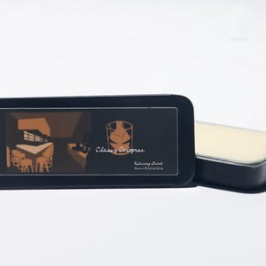 May include: A black rectangular tin with a sliding lid, featuring a graphic of a room with tables and chairs, and the text "Classy Colognes." Beside it is a rectangular solid cologne. The cologne is a light cream color.