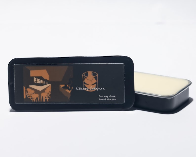 May include: A black rectangular tin with a sliding lid, featuring a graphic of a room with tables and chairs, and the text "Classy Colognes." Beside it is a rectangular solid cologne. The cologne is a light cream color.