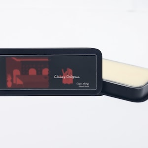 May include: A black rectangular tin with rounded corners, featuring a red graphic of a lounge scene and the text "Classy Colognes." Beside it, a matching open tin reveals a solid, light-colored cologne. The product is designed for men.
