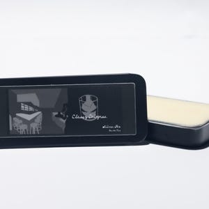 May include: A black rectangular tin with a clear lid, featuring the text "Classy Colognes" and a graphic. Beside it is a matching black tin with a light beige solid cologne inside. The background is white.