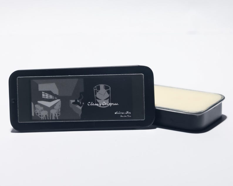 May include: A black rectangular tin with a clear lid, featuring the text "Classy Colognes" and a graphic. Beside it is a matching black tin with a light beige solid cologne inside. The background is white.