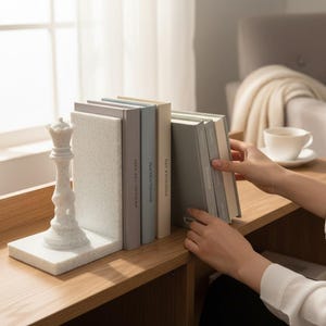 May include: A white marble queen chess piece bookend supports a row of books in various colors. The books are on a wooden shelf, with a window and a chair in the background. The scene is well-lit and has a minimalist aesthetic.